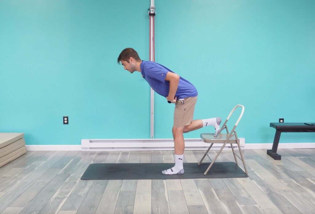 Dr. Jon is demonstrating a rear foot elevated RDL with his right foot placed on a chair standing on his left leg and hinging at both hips