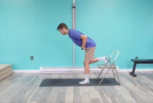 Dr. Jon is demonstrating a rear foot elevated RDL with his right foot placed on a chair standing on his left leg and hinging at both hips