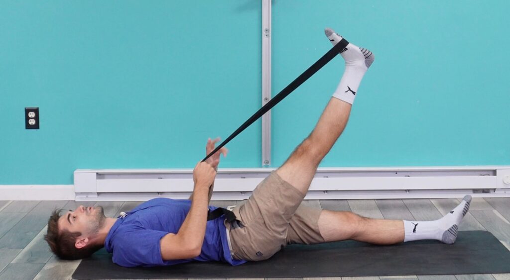 Dr. Jon deomonstrating a supine hamstring stretch lying on his back on the ground with his right leg lifted using a strap left leg straight on the ground.