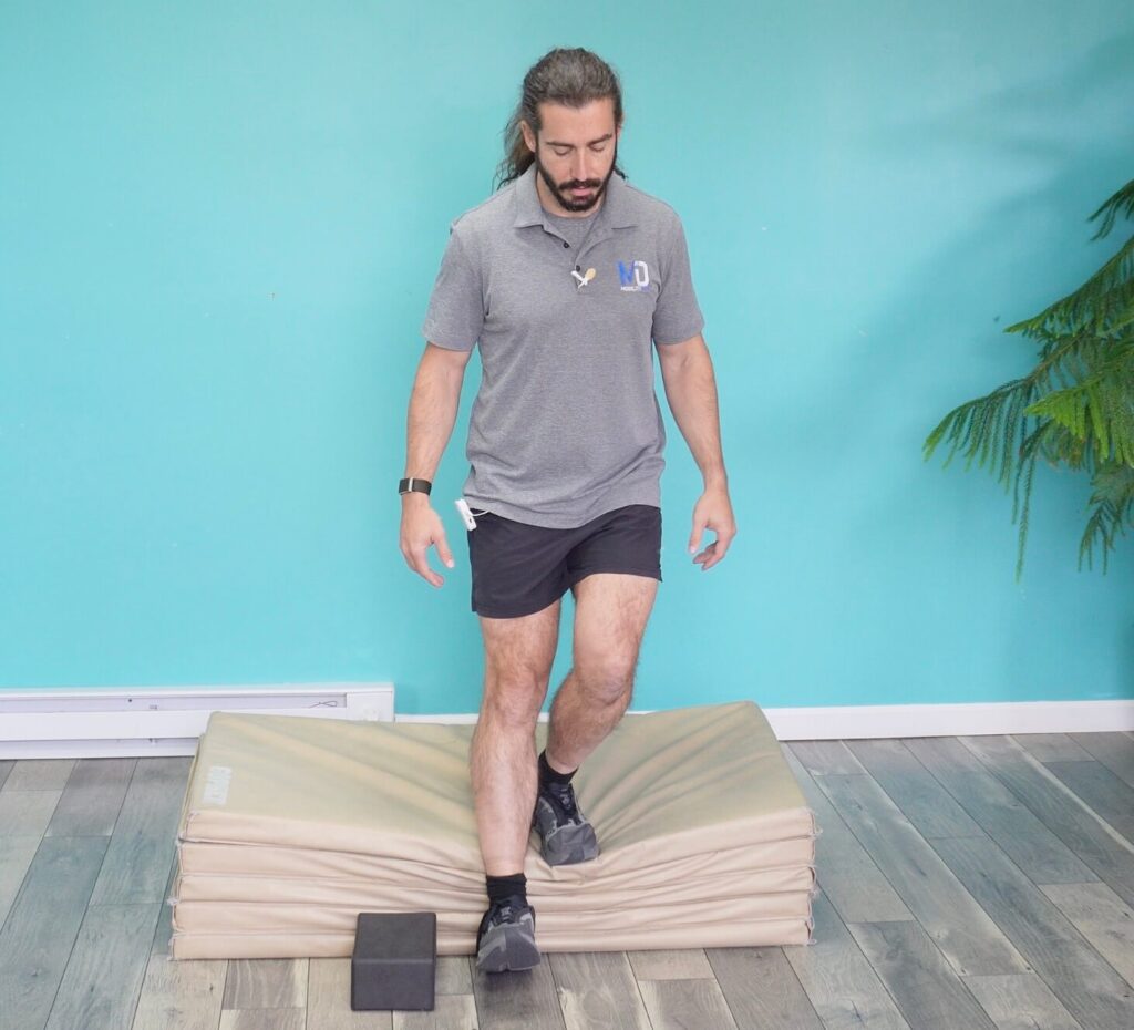 Dr. John demonstrating the standard step down exercise by standing on a mat, balancing on his left foot and lowering his right heel to touch the ground