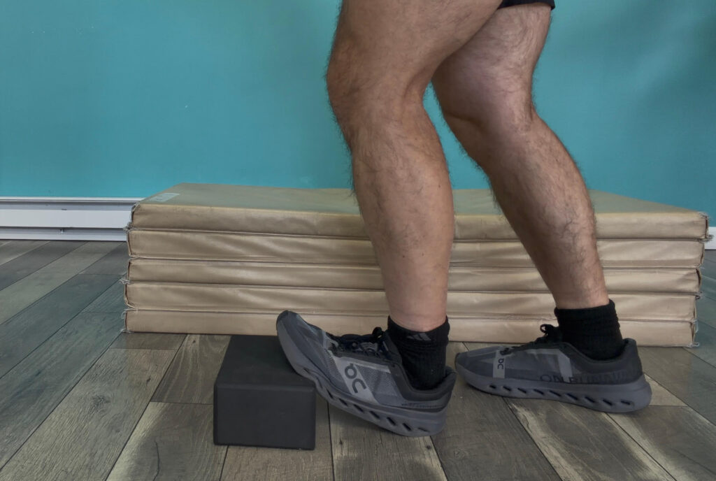 Dr. John standing in a split stance with the ball of his left foot propped up on a yoga block doing an ankle dorsiflexion stretch