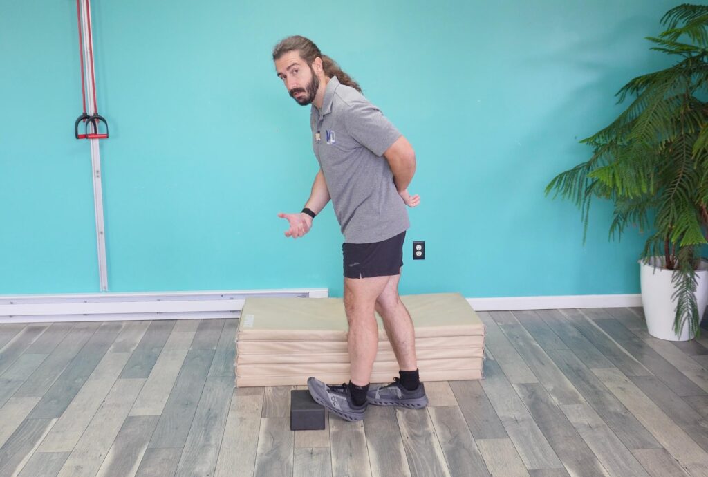 Dr. John demonstrating the straight leg ankle dorsiflexion stretch by keeping his toes up on a yoga block, the leg straight and leaning his body forward over the leg