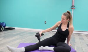 Dr. Chloe demonstrating how to use a massage gun on her quadricep - she is sitting on the ground with one leg out to the side and using the massage gun just above the knee in her quad.