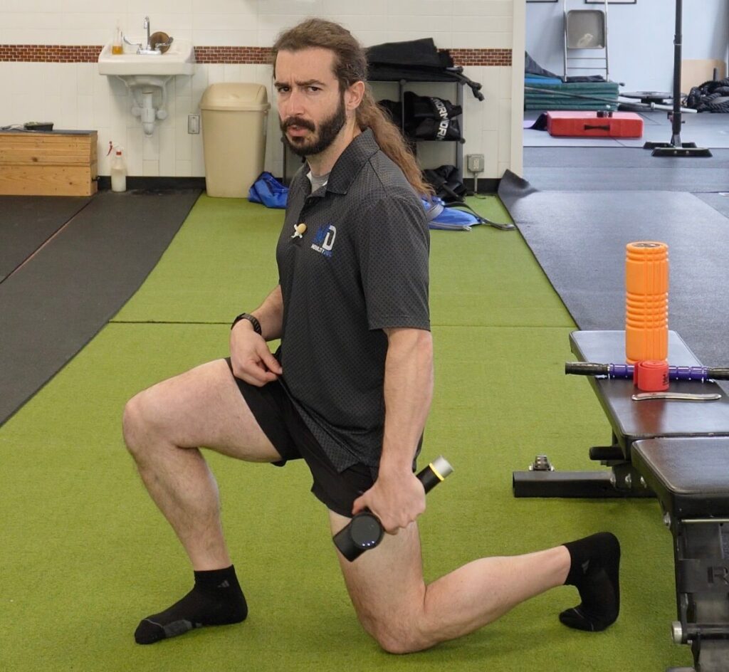 Dr. John in a kneeling hip flexor stretch using the massage gun on his extended hip flexor